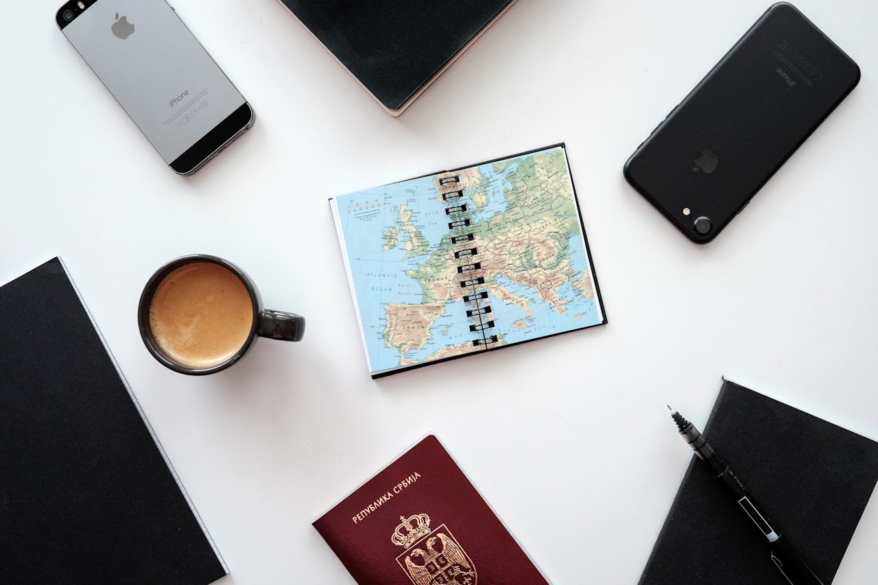 Flat lay of travel essentials with map, passport, and devices on a white surface.